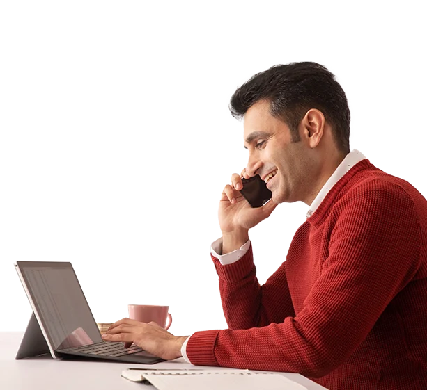 Man in a rust sweater smiles on a phone call while typing on a laptop, coffee mug beside him, orange background.