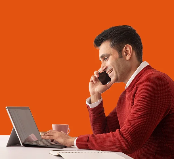 A smiling man in a red sweater talks on a mobile phone while sitting in front of a laptop.