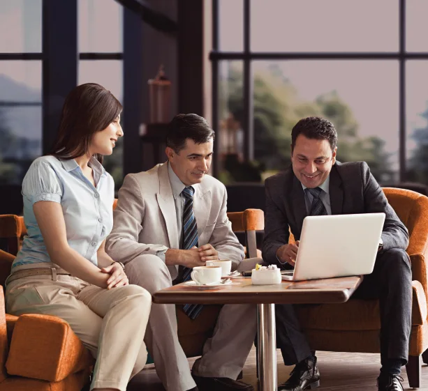 Three business professionals in formal attire sit at a table looking at a laptop screen together.