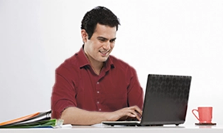 Man in red shirt working on laptop at desk with coffee mug beside him.